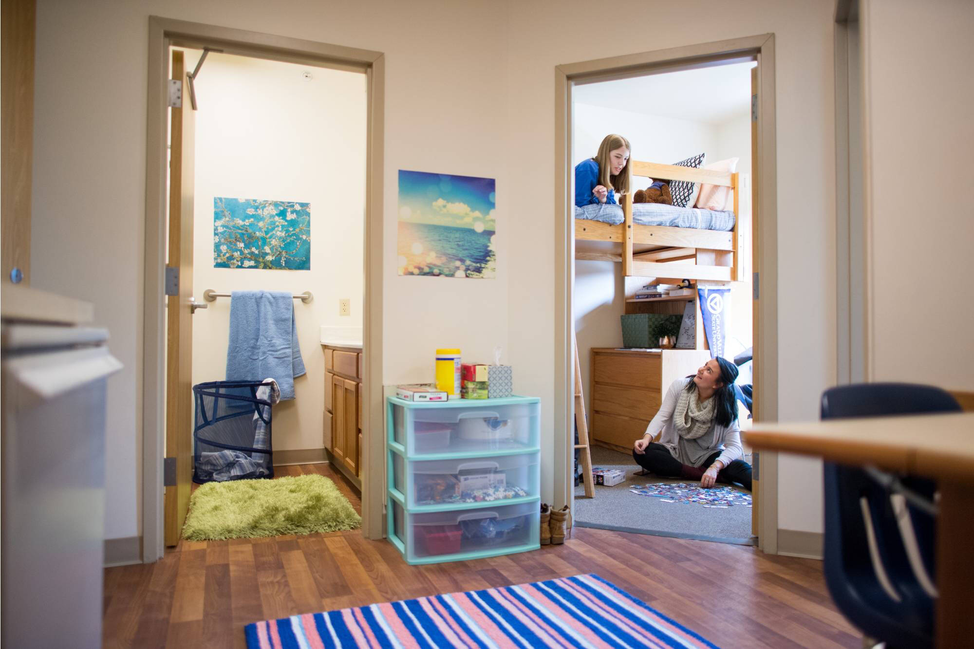 A Johnson living unit with wood flooring features a bathroom on the left and a bedroom on the right. Two people are interacting in the bedroom—one on a loft bed and the other sitting on the floor with puzzle pieces.